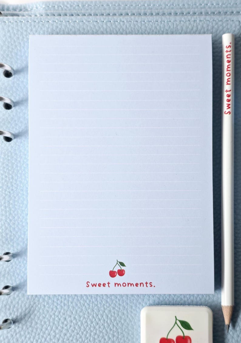 May include: A lined notepad with a cherry illustration and the text "Sweet moments." A white pencil with the text "Sweet moments." is next to the notepad. A small eraser with a cherry illustration is also present.
