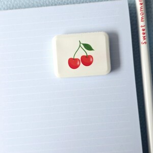 May include: A rectangular eraser with a cherry design, featuring two red cherries and a green leaf. The eraser is on a lined paper, with a pencil that reads "Sweet moments."