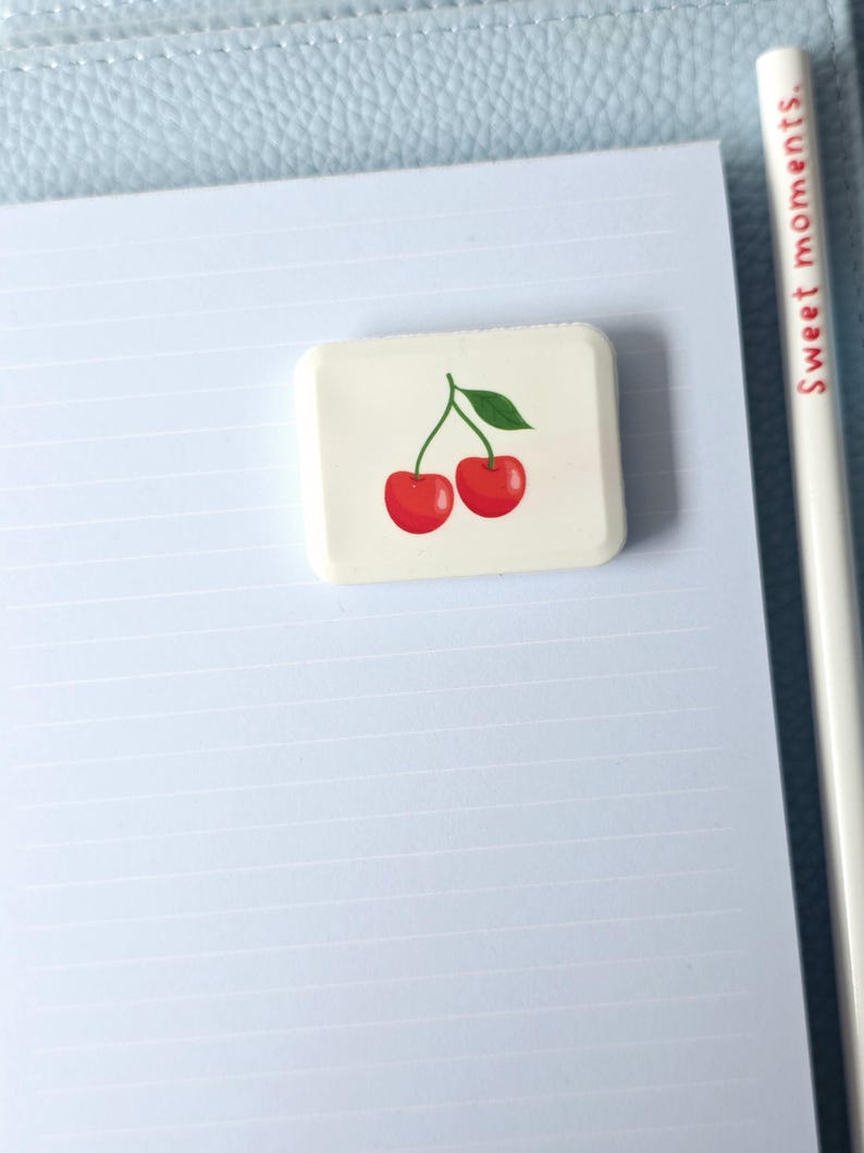 May include: A rectangular eraser with a cherry design, featuring two red cherries and a green leaf. The eraser is on a lined paper, with a pencil that reads "Sweet moments."
