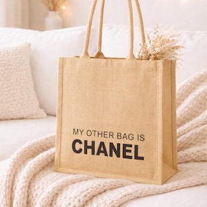 Könnte beinhalten: Beige Tragetasche mit dem Text "MY OTHER BAG IS CHANEL" in Schwarz. Die Tasche hat zwei Henkel und ist mit Trockenblumen gefüllt. Die Tasche liegt auf einer cremefarbenen Decke.