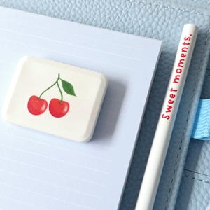 May include: A white eraser with a cherry design and a white pencil with the text "Sweet moments." are on a light blue lined notepad. The notepad is inside a light blue leather-like notebook.