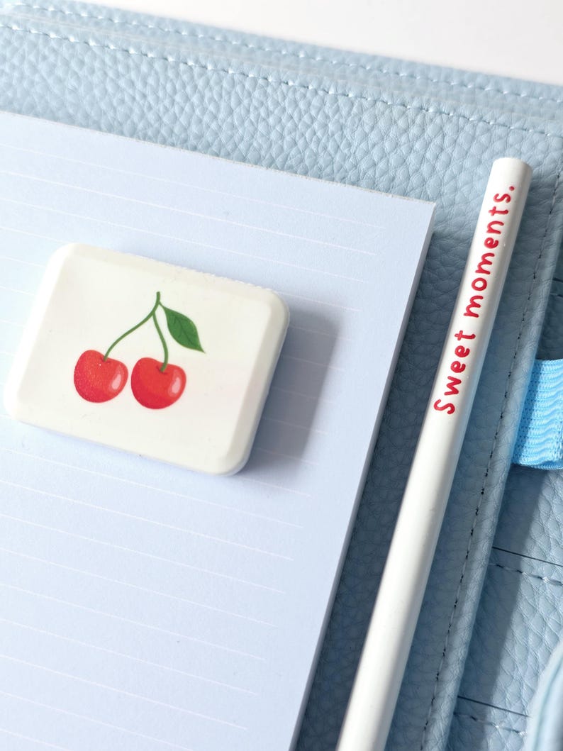 May include: A white eraser with a cherry design and a white pencil with the text "Sweet moments." are on a light blue lined notepad. The notepad is inside a light blue leather-like notebook.
