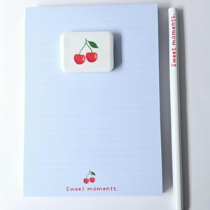 May include: A light blue notepad with horizontal lines, a white pencil, and a white square eraser. The eraser and notepad feature a cherry design. The pencil has the text "Sweet moments." in red. The notepad also has the text "Sweet moments." in red.