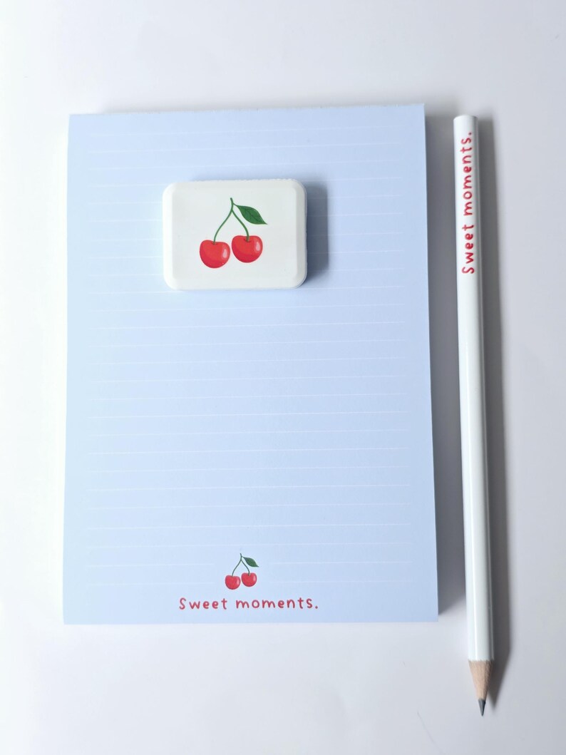 May include: A light blue notepad with horizontal lines, a white pencil, and a white square eraser. The eraser and notepad feature a cherry design. The pencil has the text "Sweet moments." in red. The notepad also has the text "Sweet moments." in red.