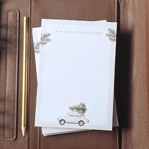 Könnte beinhalten: Ein weißer Notizblock mit dem aufgedruckten Text "WINTER WONDERLAND" oben, verziert mit Tannenzweigen in den Ecken und einem kleinen Auto mit Tannenbaum unten, begleitet von einem goldenen Bleistift mit Text an der Seite.