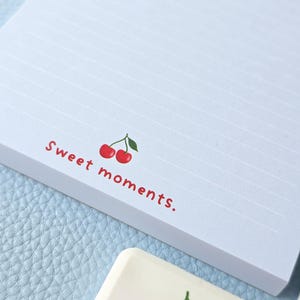 May include: A white notepad with horizontal lines and the words "Sweet moments." in red, along with a cherry illustration. A matching cherry-themed eraser is also visible. The notepad and eraser rest on a light blue surface.