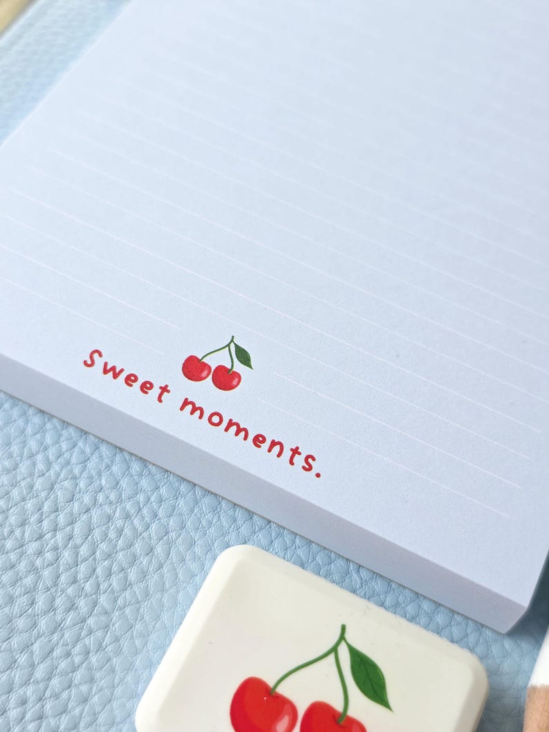 May include: A white notepad with horizontal lines and the words "Sweet moments." in red, along with a cherry illustration. A matching cherry-themed eraser is also visible. The notepad and eraser rest on a light blue surface.