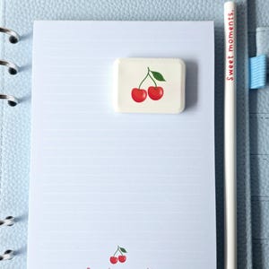 May include: A light blue planner with a lined notepad, a white pencil, and a rectangular eraser. The eraser features a red cherry design. The pencil reads "Sweet moments.", and the notepad has a cherry graphic and the text "Sweet moments."
