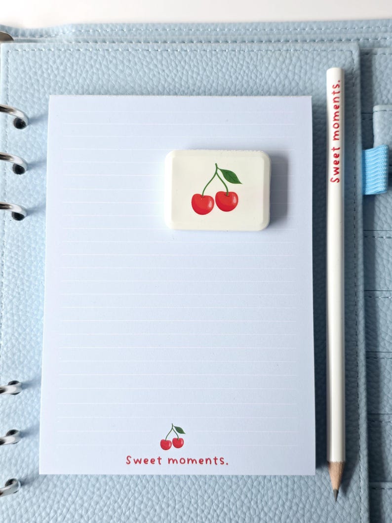 May include: A light blue planner with a lined notepad, a white pencil, and a rectangular eraser. The eraser features a red cherry design. The pencil reads "Sweet moments.", and the notepad has a cherry graphic and the text "Sweet moments."