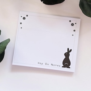 Könnte beinhalten: Weißer Notizblock mit einer schwarzen Kaninchensilhouette und dem Text "Hop On Notes". Der Notizblock hat einen gepunkteten Rand und sternförmige Designs. Ein charmantes Design zum Notieren von Notizen.