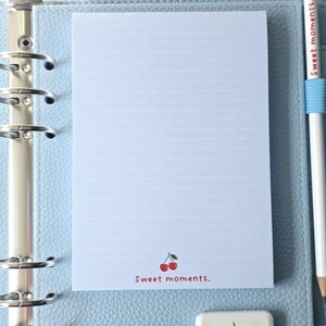 May include: A light blue binder with a lined notepad featuring a cherry illustration and the text "Sweet moments." A white pencil with the same text and a cherry eraser are also present. The binder is open, revealing the notepad.
