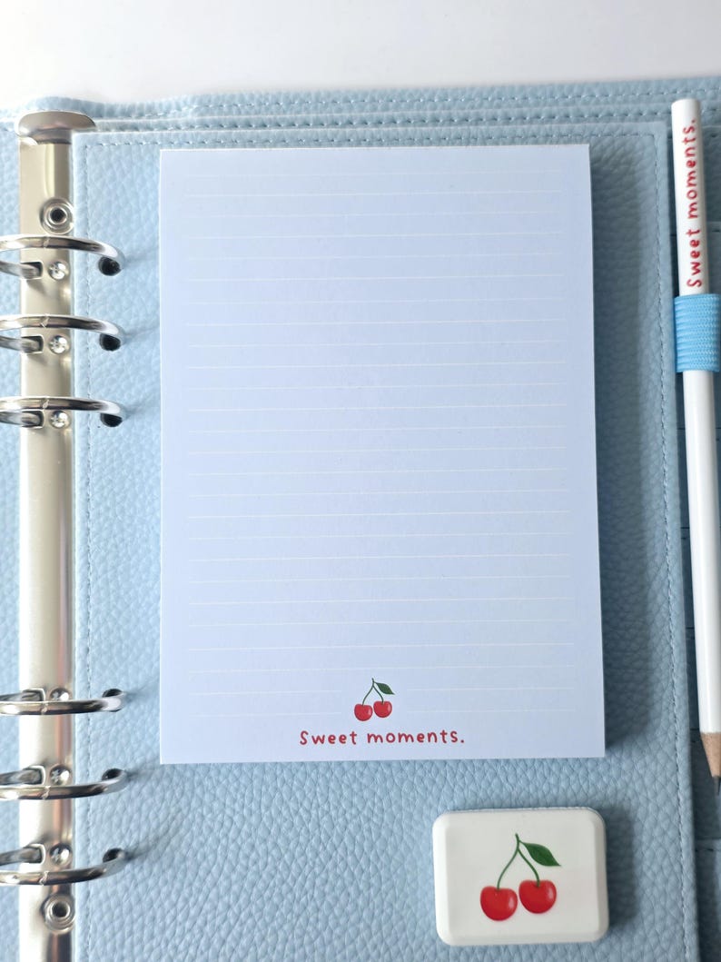 May include: A light blue binder with a lined notepad featuring a cherry illustration and the text "Sweet moments." A white pencil with the same text and a cherry eraser are also present. The binder is open, revealing the notepad.