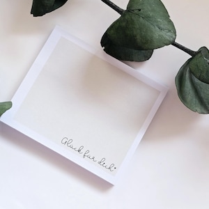 Könnte beinhalten: Eine quadratische, cremefarbene Karte mit weißem Rand, die den handgeschriebenen Satz "Glück für dich" in schwarzer Schrift zeigt. Ein Zweig mit grünen Blättern befindet sich in der oberen rechten Ecke. Die Karte liegt auf einem weißen Hintergrund.