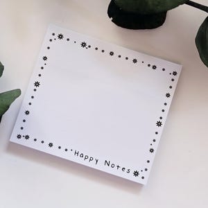 Könnte beinhalten: Ein weißer Notizblock mit einem Rand aus kleinen schwarzen Stern- und Punktmustern. Die Worte "Happy Notes" sind unten in Schwarz gedruckt. Der Notizblock liegt auf einer weißen Oberfläche mit grünen Blättern im Hintergrund.