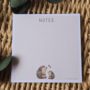 Könnte beinhalten: Ein quadratischer Notizblock mit dem Wort „NOTES“ oben. Der Notizblock hat einen hellbeigen Hintergrund mit kleinen Punkten und eine Illustration von zwei Igeln mit Herzen darüber. Der Notizblock liegt auf einer gewebten Oberfläche.