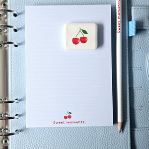 May include: A light blue planner with a lined notepad, a white pencil, and a cherry-themed eraser. The notepad has the words "Sweet moments." printed on it. The pencil has the words "Sweet moments." printed on it.