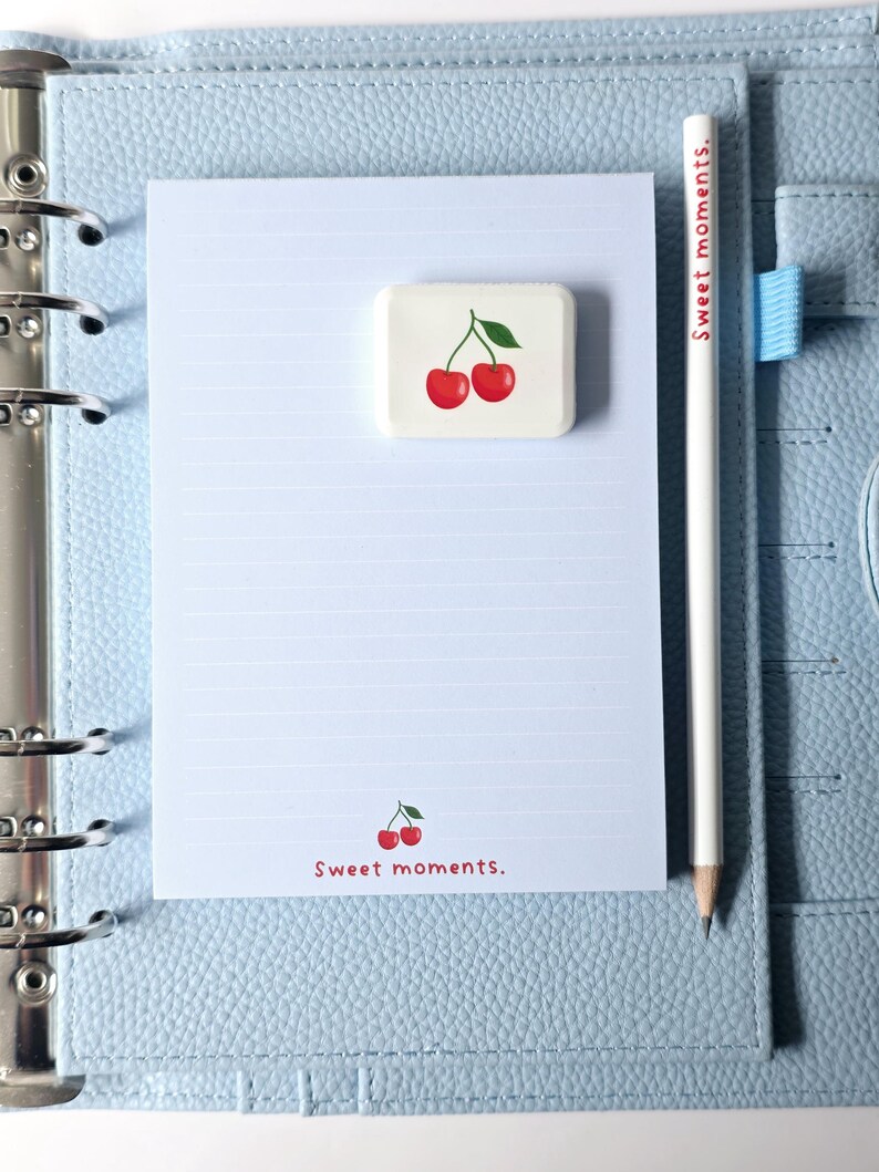May include: A light blue planner with a lined notepad, a white pencil, and a cherry-themed eraser. The notepad has the words "Sweet moments." printed on it. The pencil has the words "Sweet moments." printed on it.