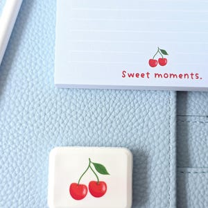 May include: A light blue notepad and a white rectangular eraser with a cherry design. The notepad has horizontal lines and the text "Sweet moments." The eraser features two red cherries with a green leaf.