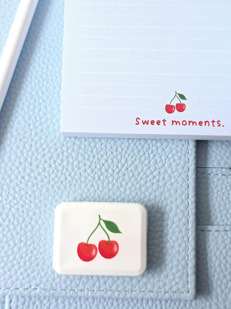 May include: A light blue notepad and a white rectangular eraser with a cherry design. The notepad has horizontal lines and the text "Sweet moments." The eraser features two red cherries with a green leaf.