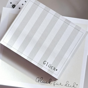 Könnte beinhalten: Ein Stapel weiß-grau gestreifter Notizkarten mit dem Wort "Glück" und "Glück für dich" in schwarzer Schrift. Die oberste Karte hat einen gepunkteten Rand. Andere Karten haben Blumen- und Sterndesigns.