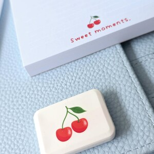May include: A white rectangular eraser with a cherry design and a notepad with the text "Sweet moments." The eraser and notepad are on a light blue leather surface.