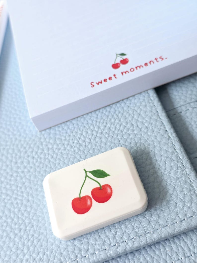 May include: A white rectangular eraser with a cherry design and a notepad with the text "Sweet moments." The eraser and notepad are on a light blue leather surface.