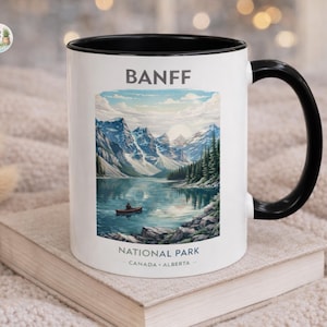 May include: White ceramic mug with a black interior and handle. The mug features a scenic illustration of Banff National Park, with text that reads "BANFF" and "NATIONAL PARK CANADA ALBERTA". The image depicts a lake surrounded by mountains.
