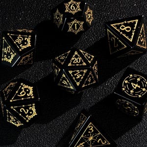 May include: A set of seven black polyhedral dice with gold-colored numbering and decorative accents. The dice are in various geometric shapes, including a d4, d6, d8, d10, d12, d20, and a d100. The dice are arranged on a dark surface.
