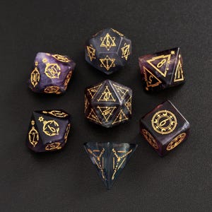 Puede incluir: Conjunto de siete dados poliédricos en tonos morados y negros con marcas doradas. Los dados tienen varias formas geométricas, incluyendo un d4, d6, d8, d10, d12, d20 y un d100. Los dados están dispuestos sobre una superficie oscura.