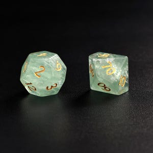 May include: Two translucent green dice with gold numbering are displayed against a black background. One die is a 12-sided d12, and the other is a 10-sided d10. The dice are made of a material that appears to have internal veining.