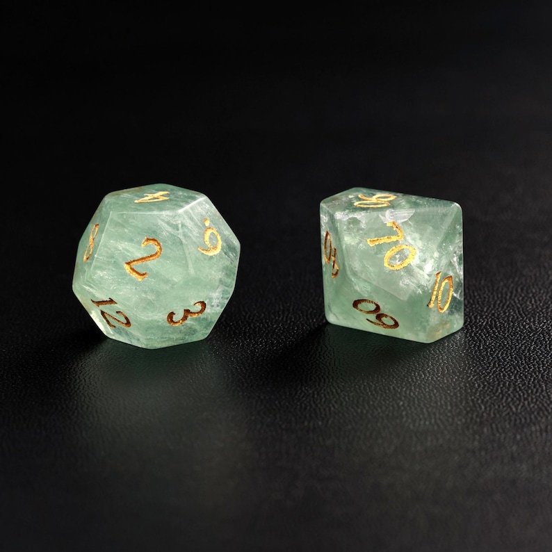 May include: Two translucent green dice with gold numbering are displayed against a black background. One die is a 12-sided d12, and the other is a 10-sided d10. The dice are made of a material that appears to have internal veining.