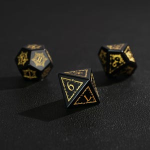 May include: Three black polyhedral dice with gold numbering and decorative accents. The dice are in various shapes, including a d20, d10, and d6. The dice are for tabletop games.
