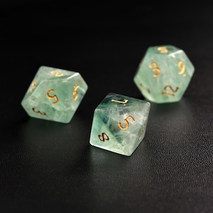 May include: Three translucent green dice with gold-colored numbers are displayed against a dark background. The dice are multi-sided, with visible internal patterns. The numbers are clearly visible, and the overall aesthetic is elegant and detailed.