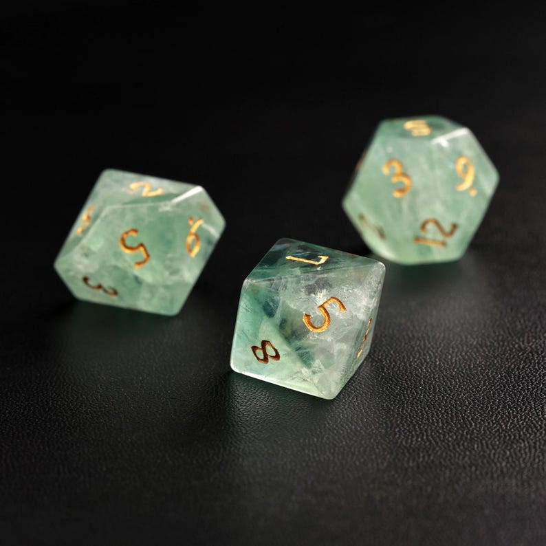May include: Three translucent green dice with gold-colored numbers are displayed against a dark background. The dice are multi-sided, with visible internal patterns. The numbers are clearly visible, and the overall aesthetic is elegant and detailed.