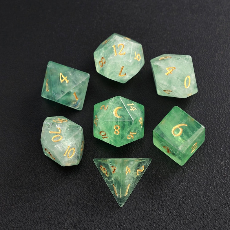May include: Set of seven green polyhedral dice with gold numbering. The dice are made of a translucent material, and the numbers are clearly visible. The dice are arranged on a black surface.