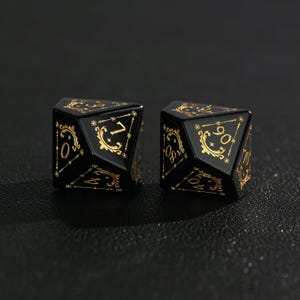 May include: Two black, multi-sided dice with gold-colored numbers and decorative flourishes. The dice are designed for role-playing games. The numbers are in a gold color and are set against a black background.