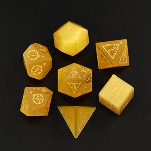 May include: Set of seven yellow gemstone dice with gold accents and etched numbers. The dice include a d4, d6, d8, d10, d%, d12, and d20.
