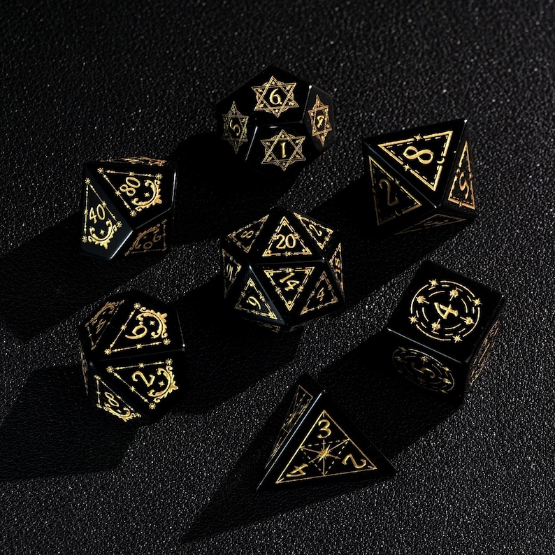 May include: A set of seven black polyhedral dice with gold-colored numbering and decorative accents. The dice are in various geometric shapes, including a d4, d6, d8, d10, d12, d20, and a d100. The dice are arranged on a dark surface.
