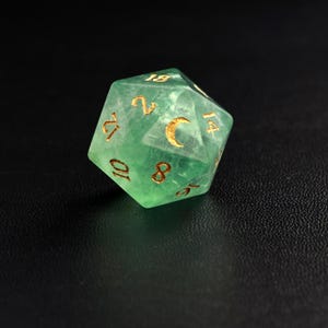 May include: A translucent green, twenty-sided die with gold-colored numbers and a crescent moon symbol. The die is designed for role-playing games and is set against a dark background.
