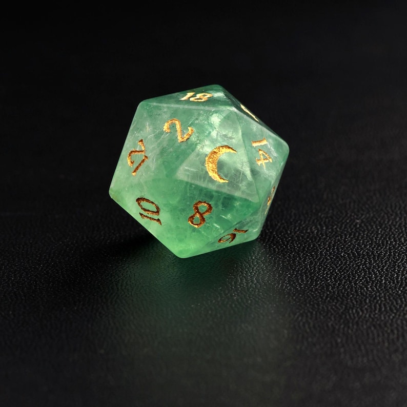 May include: A translucent green, twenty-sided die with gold-colored numbers and a crescent moon symbol. The die is designed for role-playing games and is set against a dark background.