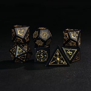 May include: A set of seven black polyhedral dice with gold-colored numbering and decorative accents. The dice include a D4, D6, D8, D10, D12, D20, and a percentile die, suitable for tabletop role-playing games.