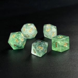May include: Five green gemstone dice with gold numbering are arranged on a dark surface. The dice are various shapes, including a d20, d12, d8, and d6, and are used for tabletop role-playing games.