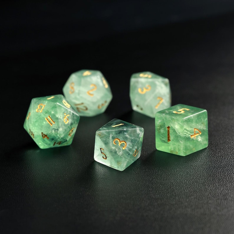 May include: Five green gemstone dice with gold numbering are arranged on a dark surface. The dice are various shapes, including a d20, d12, d8, and d6, and are used for tabletop role-playing games.