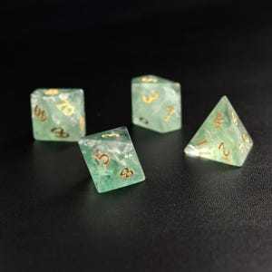 May include: Four translucent green dice with gold numbering are arranged on a black surface. The dice include a D4, a D6, and two D10s. The dice are made of a marbled, semi-precious material.