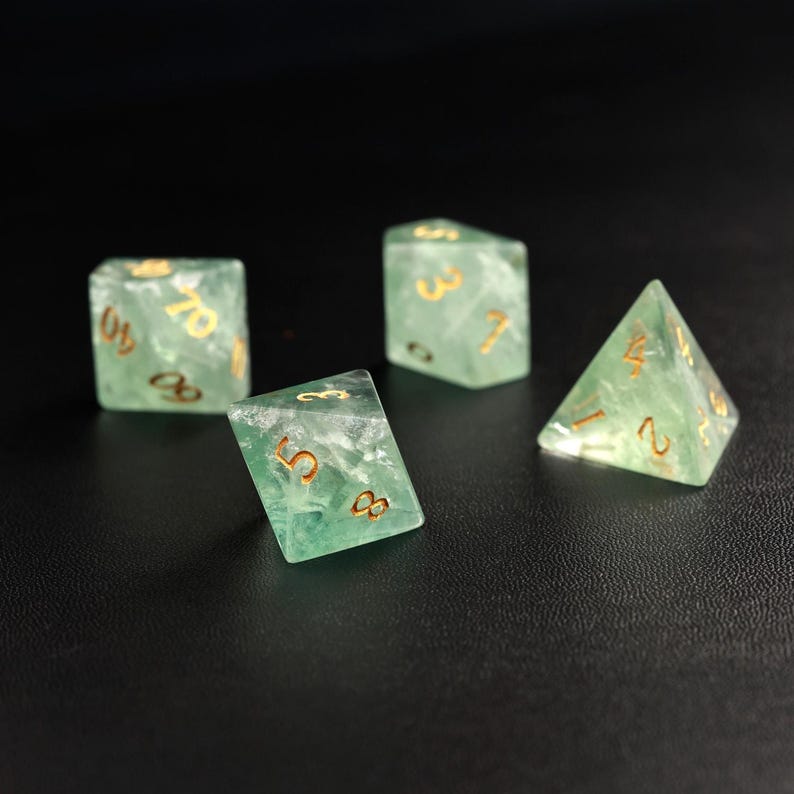 May include: Four translucent green dice with gold numbering are arranged on a black surface. The dice include a D4, a D6, and two D10s. The dice are made of a marbled, semi-precious material.