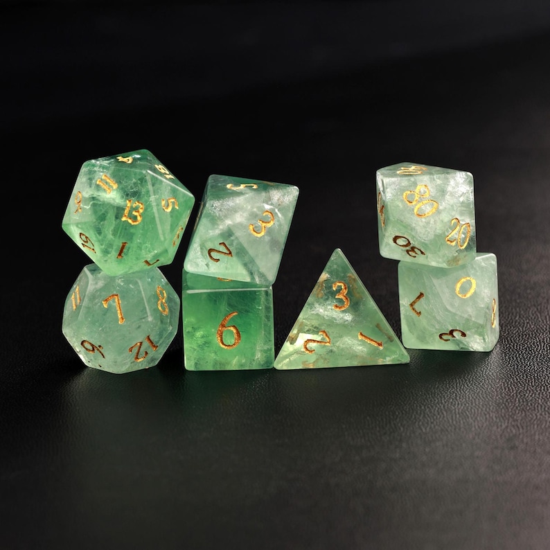 May include: A set of seven translucent green dice with gold numbering. The dice are in various shapes, including a d20, d12, d10, d8, d6, d4, and a d10 percentile. The dice are arranged on a dark surface.