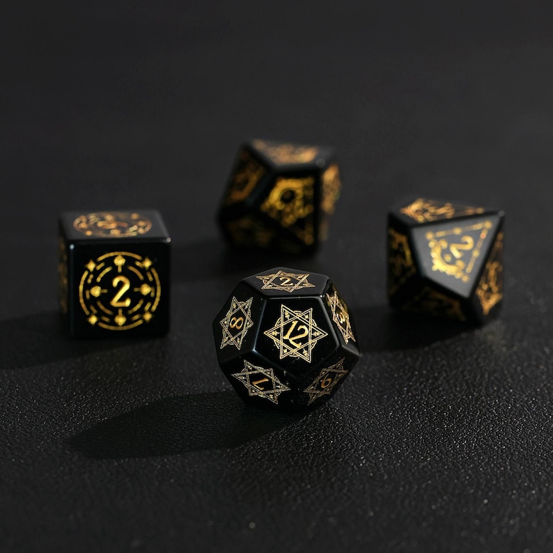 May include: Four black polyhedral dice with gold-colored markings. The dice include a six-sided die, a twenty-sided die, and two other dice with different numbers of sides. The dice are for tabletop games.