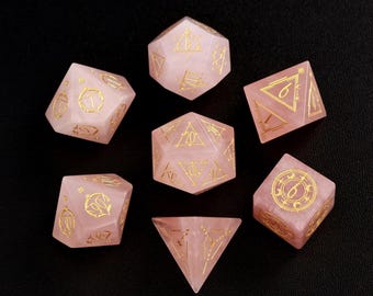 May include: Set of seven pink rose quartz dice with gold metallic numbers and symbols. The dice are in the shape of a d4, d6, d8, d10, d%, d12, and d20.