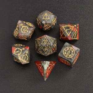 Op de afbeelding: Set van zeven polyhedrische dobbelstenen gemaakt van rood, zwart en groen steen met gouden accenten. De dobbelstenen zijn genummerd 4, 6, 8, 12, 20 en een d4.