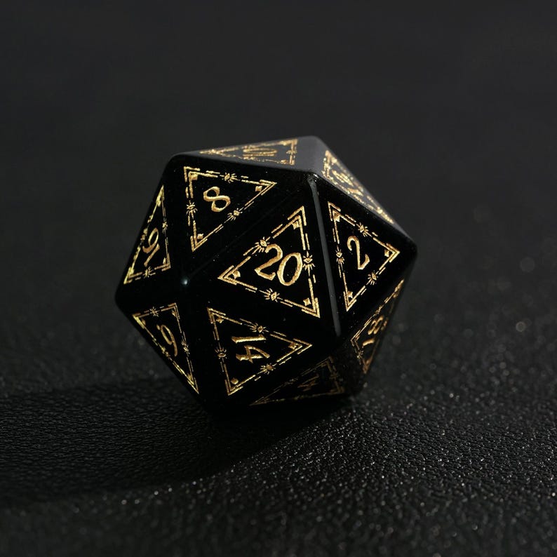 May include: A black twenty-sided die with gold-colored numbers and decorative trim. The die is a polyhedron, with each face displaying a number from 1 to 20. The numbers are in a serif font.